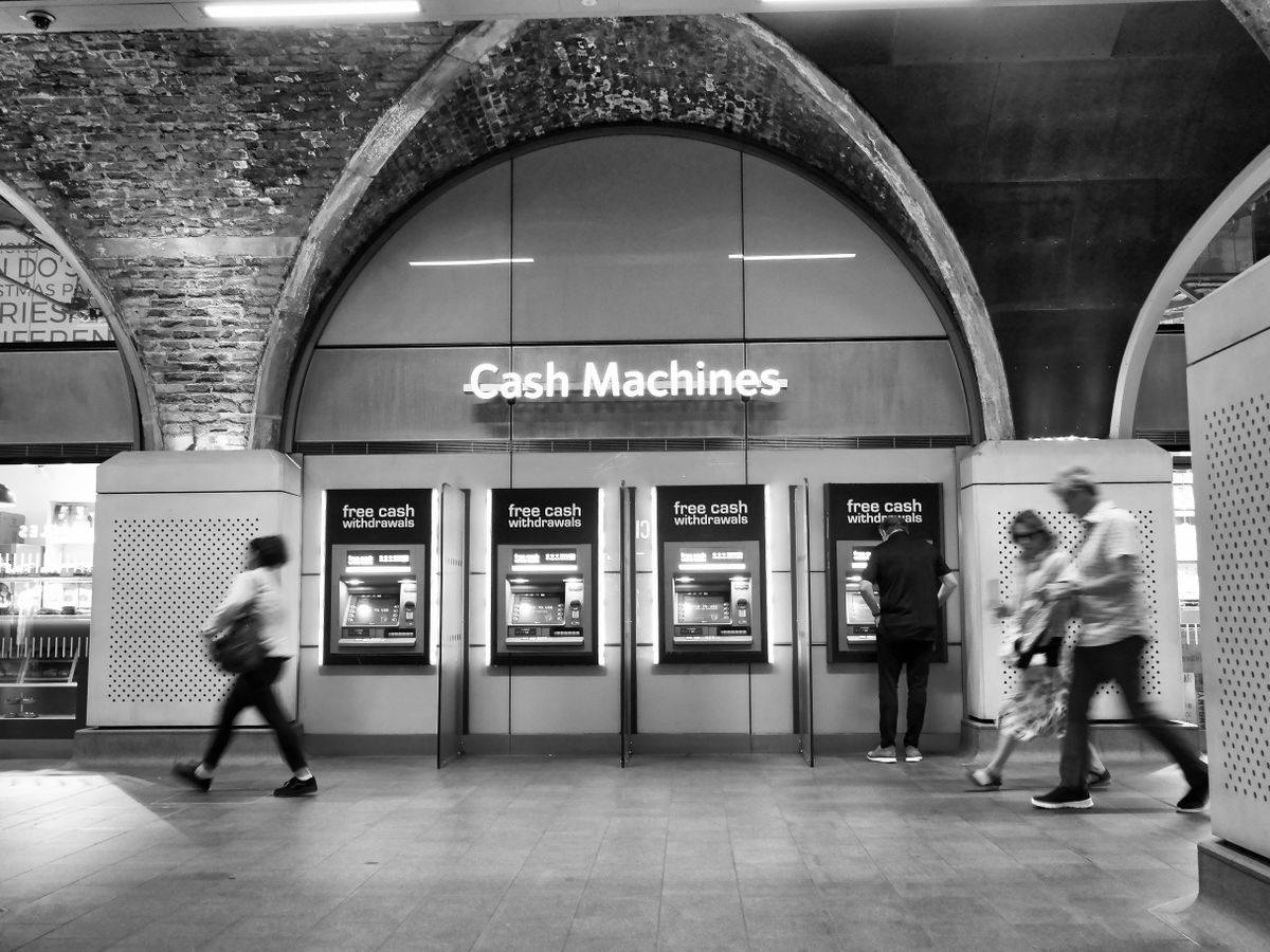 Pessoas usando caixas eletrônicos em uma estação, em preto e branco.